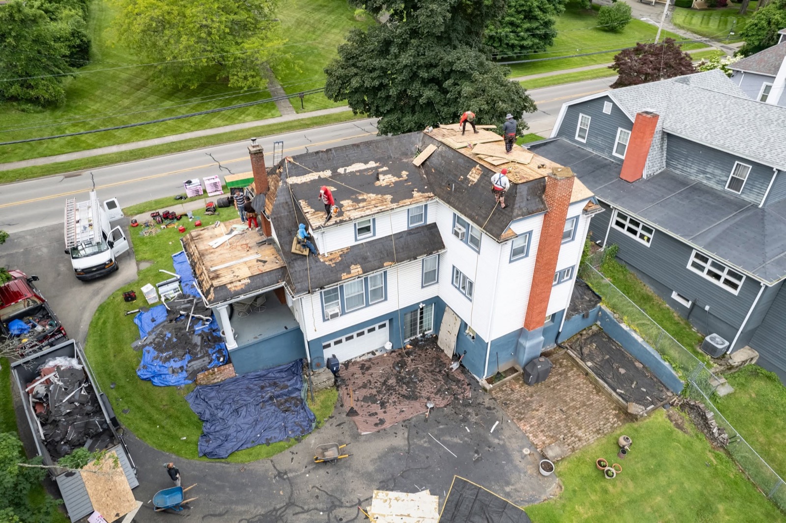 Drone view of the Rail Roofing Group crew mid tear-off on a large home, tarps and dump trailer staged below
